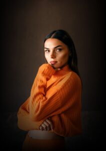 Young woman wearing an oversized orange ribbed sweater, posing confidently against a dark background with soft studio lighting. The fashion portrait highlights a modern, elegant, and minimalist style.