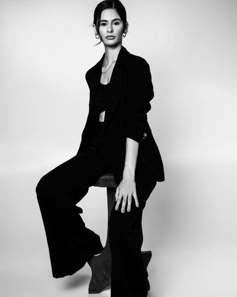 Black-and-white portrait of a fashion model wearing a tailored black blazer and wide-leg trousers, seated on a wooden stool in a minimalist studio setting.